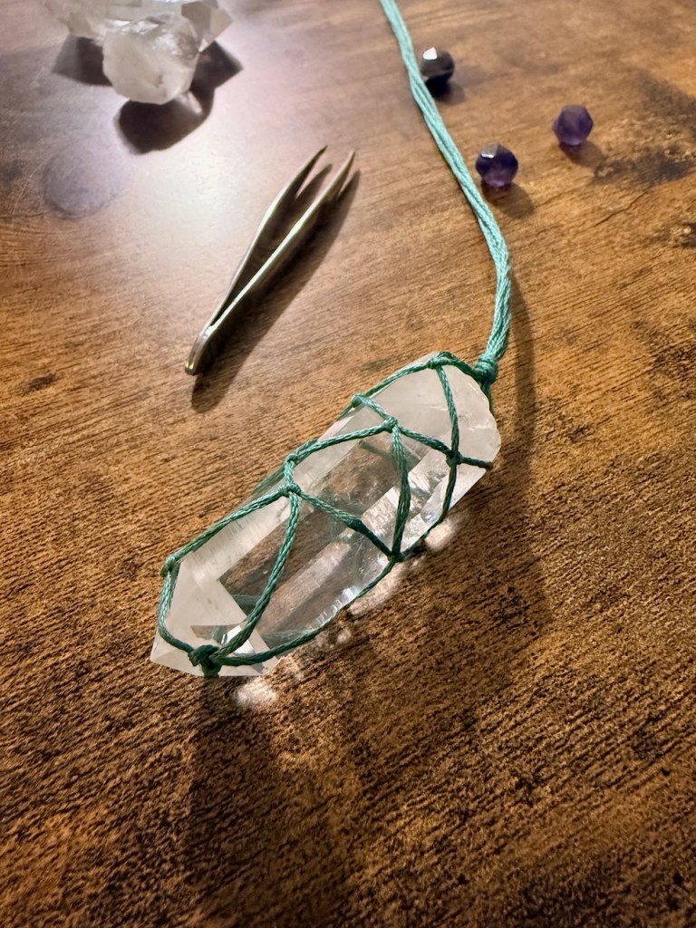 a clear quartz point, wrapped in a green macrame net, lying on a brown desk with a pair of tweezers and small beads in the background