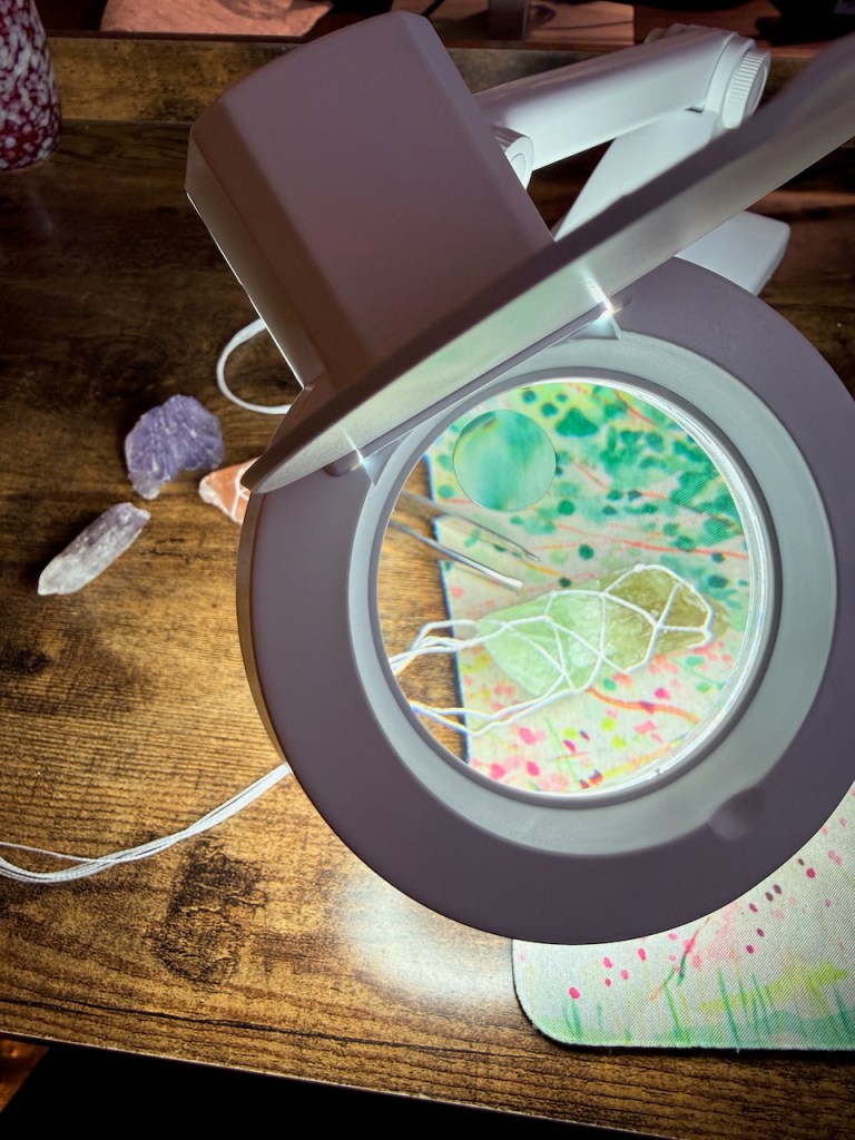 a piece of green flourite being wrapped in a white thread macrame net, seen through a magnifying lamp