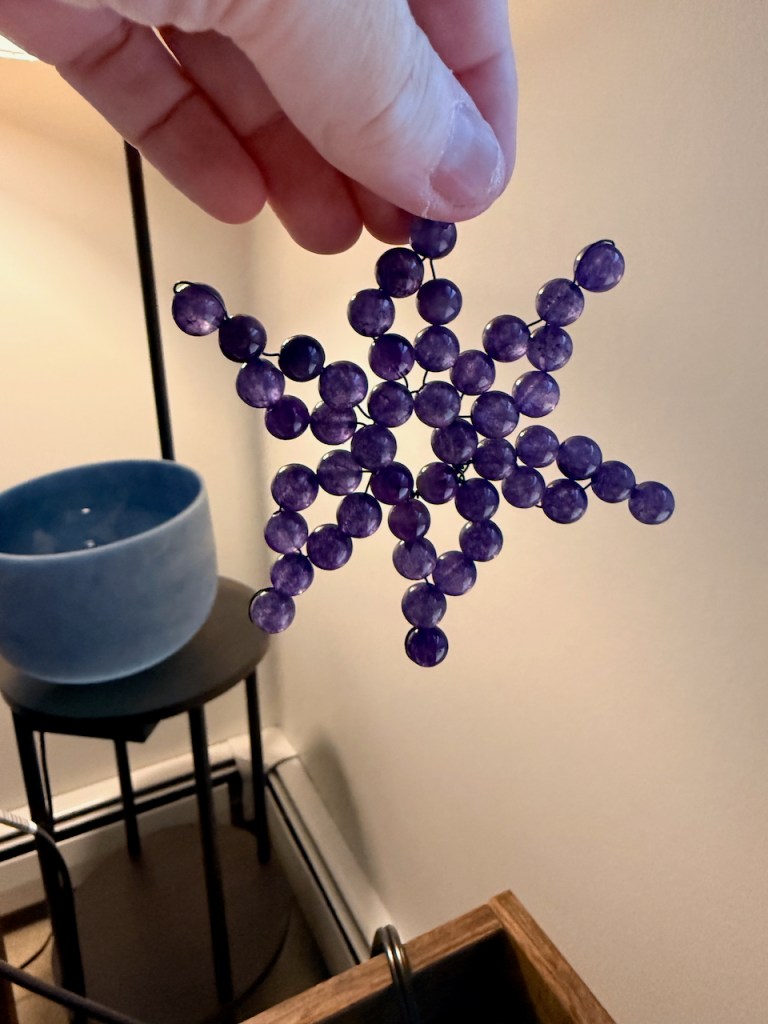 fingers holding a snowflake-shaped ornament made from round purple amethyst beads